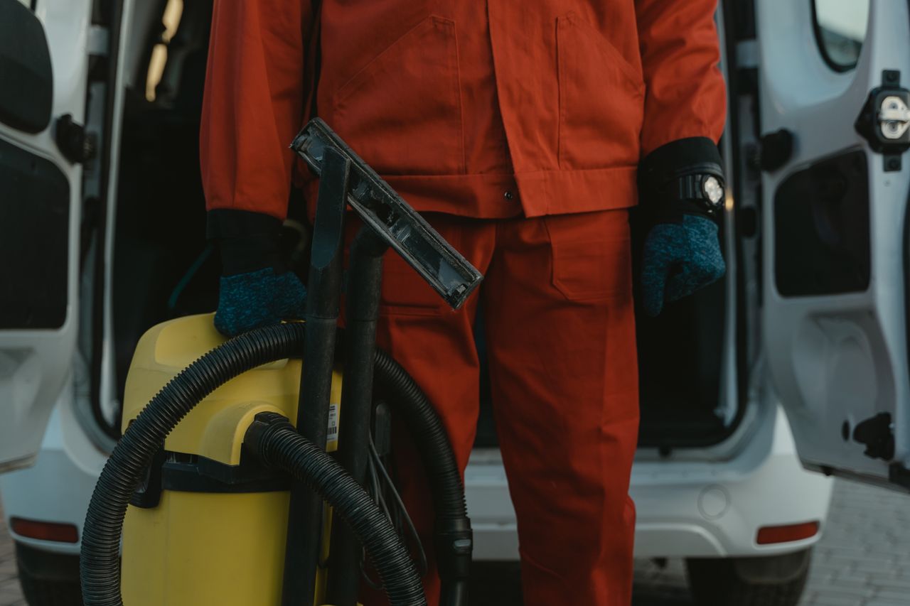 Une professionnelle du nettoyage en uniforme rouge tenant un aspirateur près d'un van, prête à intervenir.